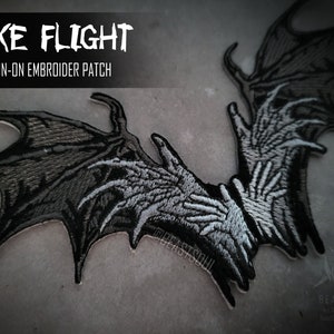 May include: An iron-on embroidered patch with a dark, gothic design. The patch features a pair of stylized wings with a skeletal hand design in shades of gray and black. Text at the top reads "TAKE FLIGHT" and "IRON-ON EMBROIDER PATCH".