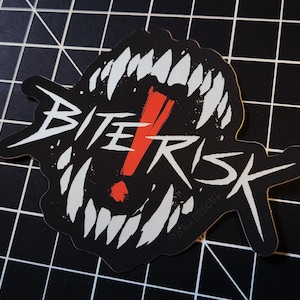 May include: A black sticker with the words "BITE RISK" in white, stylized to resemble a mouth full of sharp teeth. A red exclamation point is in the center. The sticker is on a black surface with a white grid pattern.