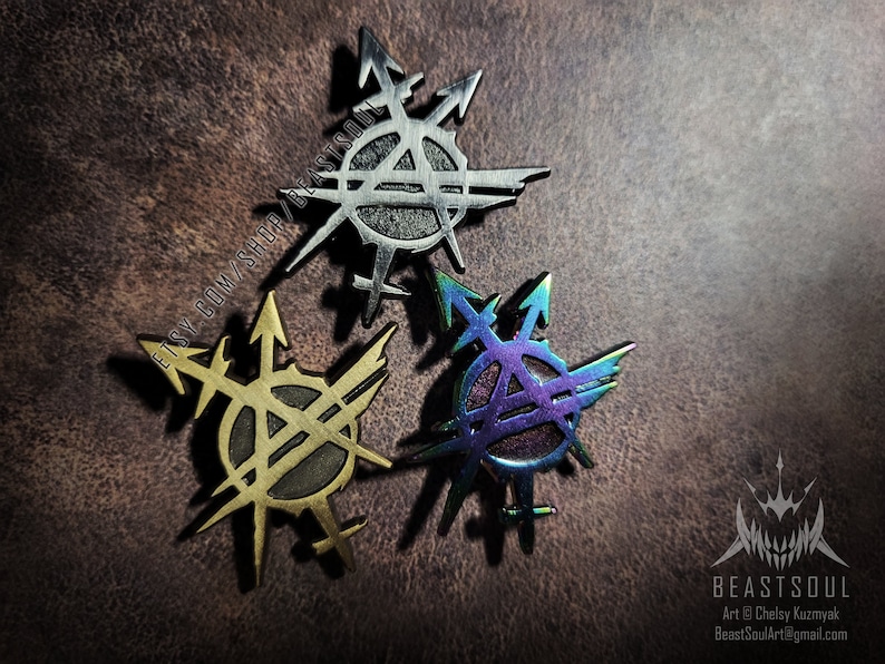 K&ouml;nnte beinhalten: Drei Metallnadeln mit einem stilisierten Design mit Pfeilen und einem Kreis mit dem Buchstaben "A" darin. Eine Nadel ist silberfarben, eine ist goldfarben und eine hat ein regenbogenfarbenes, schillerndes Finish. Die Nadeln liegen auf einer strukturierten braunen Oberfl&auml;che.
