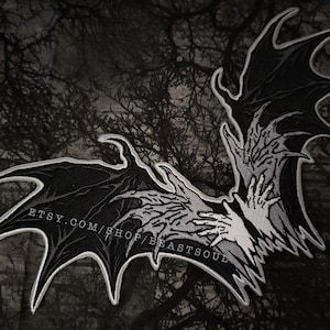 May include: Black and white embroidered patch of two bat wings with skeletal hands reaching out from the centre. The patch has a white outline and is on a dark background.
