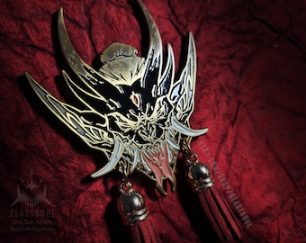 May include: A detailed, gold-toned metal pin featuring a stylized demon face with a red eye and intricate black and gold detailing. The pin has dangling tassels and a small bell. The background is a textured, deep red fabric.