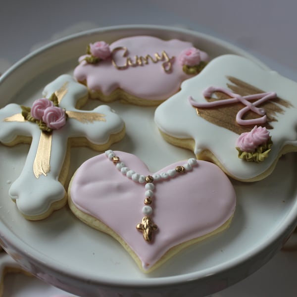 Könnte beinhalten: Ein Set dekorierter Zuckerkekse mit rosa und weißem Zuckerguss. Die Kekse sind in Form eines Kreuzes, eines Herzens und eines Rechtecks. Das Kreuz hat goldene Akzente und eine rosa Blume. Das Herz hat eine weiße Rosenkranz und ein goldenes Kreuz. Das Rechteck hat einen goldenen Pinselstrich und eine rosa Blume. Die Kekse befinden sich auf einem weißen Teller.