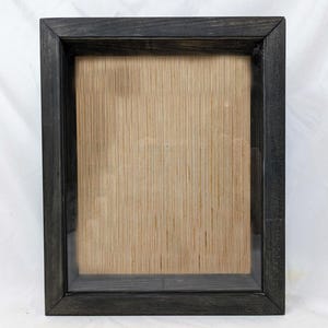 May include: A black wooden shadow box frame with a clear glass front and a light brown backing.