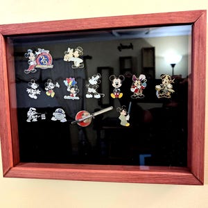 May include: A framed display case with a black background and glass front, showcasing a collection of 16 Disney character pins. The pins feature Mickey Mouse, Minnie Mouse, Goofy, and other beloved characters. The frame is made of dark red wood.
