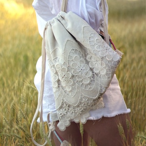 Può includere: Uno zaino beige con coulisse e dettagli in pizzo color crema. Il pizzo presenta motivi floreali e geometrici, aggiungendo un tocco delicato alla borsa. Lo zaino ha spallacci beige e un accessorio a nappa.