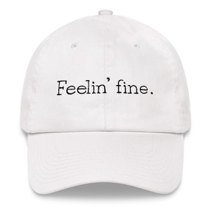 以下が含まれることがあります： 「Feelin' fine」の刺繍が入った白い野球帽。つばがカーブしており、上部にボタンが付いています。テキストはシンプルな黒いフォントです。