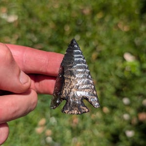 May include: A triangular arrowhead crafted from dark, translucent obsidian with a sharp, pointed tip and a notched base. The surface features distinct, rhythmic pressure-flaked ridges that create a layered, scalloped texture across the stone.