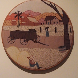 Könnte beinhalten: Ein brauner Holz-Stickrahmen mit einer bunten Stoffszene einer Farm mit einer Windmühle, einem Wagen, einem Haus und einer Person, die auf dem Feld sitzt. Der Stoff ist mit braunem, rotem, blauem und grünem Garn gestickt.