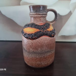 Peut inclure: Un vase en céramique brun avec un glaçage noir et orange. Le vase a un corps arrondi et une petite poignée.