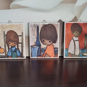 May include: Three vintage square art prints featuring illustrations of children. The prints depict children in various settings, with a muted color palette of blues, oranges, and browns. Each print has a small metal hanger.