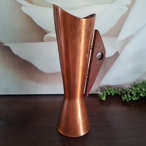 Op de afbeelding: Een koperen vaas met een houten handvat. De vaas heeft een uitlopende basis en een smalle hals. Het handvat is aan de zijkant van de vaas bevestigd en heeft een klein gaatje.