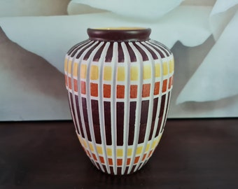 Può includere: Un vaso in ceramica marrone con un motivo geometrico di quadrati e strisce gialle, arancioni e bianche.