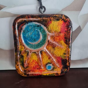 Könnte beinhalten: Quadratischer, farbenfroher Anhänger mit Kette. Das Design zeigt ein sonnenähnliches Motiv in Blau, Gelb, Orange und Rot. Der Anhänger ist an einer schwarzen Metallkette und einem Ring befestigt.
