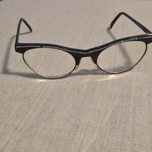 Op de afbeelding: Zwarte cat-eye bril met heldere glazen. De monturen zijn versierd met kleine, sprankelende strass-steentjes langs de bovenrand. De brug en scharnieren zijn goudkleurig, wat een contrast vormt met de donkere monturen. Op een neutrale achtergrond.