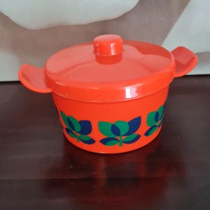 May include: An orange plastic cooking pot with a lid and two side handles. The pot has a floral pattern in blue and green. The pot is sitting on a dark brown surface.