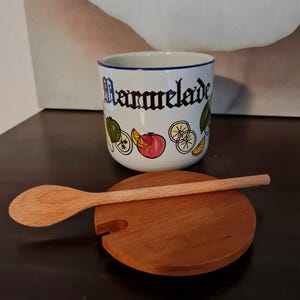 Peut inclure: Pot en céramique blanche avec un bord bleu et un couvercle et une cuillère en bois. Le pot a un motif de fruits colorés et le mot "Marmelade" écrit dessus.