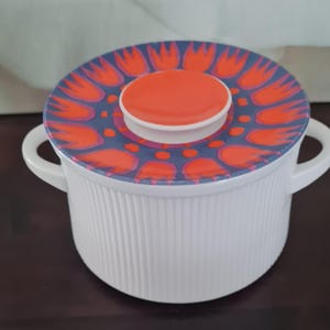 Può includere: Pirofila in ceramica bianca con coperchio con un motivo floreale arancione e viola. Il piatto ha due manici e dettagli a coste verticali. Il coperchio ha un pomello arancione.
