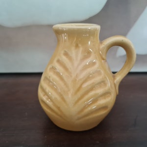 Könnte beinhalten: Ein kleiner, gelber Keramik Krug mit einem Blattdesign. Der Krug hat einen Henkel und einen runden Körper.