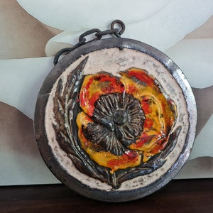 Puede incluir: Un colgante de pared redondo de cerámica con un diseño en relieve tridimensional de una flor de amapola roja, naranja y amarilla.