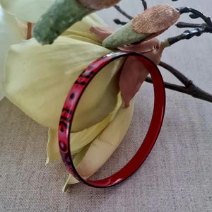 Può includere: Un braccialetto a cerchio con motivi rossi e neri. Il braccialetto ha una finitura lucida ed è posto su uno sfondo di un fiore giallo e un rametto con accenti verdi e marroni. Il braccialetto è un gioiello.