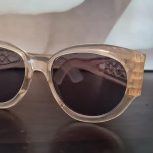 Könnte beinhalten: Cat-Eye-Sonnenbrille mit hellbraunem Rahmen und dunklen Gläsern. Der Rahmen hat ein dekoratives geometrisches Design in der Nähe der Scharniere. Die Sonnenbrille ist vor einem neutralen Hintergrund platziert.