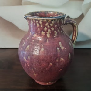 May include: A red ceramic pitcher with a speckled glaze and a handle. The pitcher has a rounded body and a slightly flared lip.