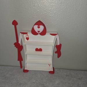 May include: A whimsical, white and red shelf unit shaped like the Queen of Hearts from Alice in Wonderland. The unit has three shelves, red heart accents, and a red scepter. The card suit symbols are visible on the shelf.