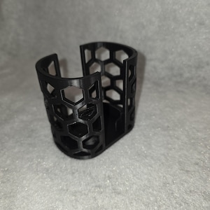 May include: A black, 3D-printed cup holder with a honeycomb pattern. The holder has a curved top and a flat base. The open design allows for easy access and visibility of the contents. The holder is made of a durable plastic material.