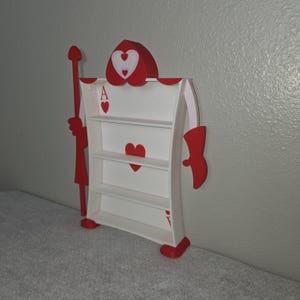 May include: A whimsical, red and white Ace of Hearts playing card-shaped bookshelf. The bookshelf has three shelves and is adorned with heart symbols. It features a red spear and decorative elements, creating a unique, decorative storage solution.