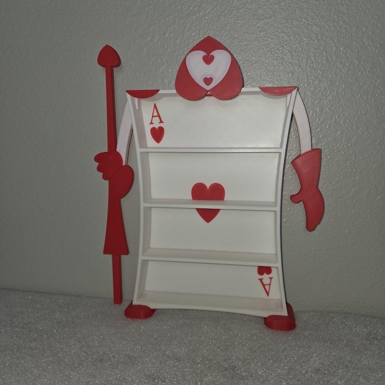 May include: A whimsical, white and red Ace of Hearts bookshelf. The bookshelf features three shelves and is decorated with red hearts and the letter "A". The design includes arms, legs, and a spear, creating a playful, decorative piece.