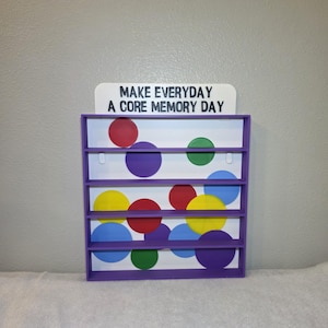 May include: A purple wooden shelf unit with five shelves. The back of the unit is white with colorful polka dots in red, purple, green, yellow, and blue. The top of the unit has a white sign that reads "MAKE EVERYDAY A CORE MEMORY DAY."