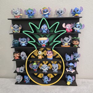 May include: A black display shelf in the shape of a pineapple, filled with various Stitch figurines. The figurines are in different poses and outfits, with blue, pink, and yellow accents. The shelf is mounted on a white wall.