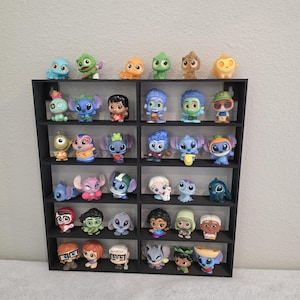 Könnte beinhalten: Ein schwarzes Regal voller bunter Charakterfiguren. Die Figuren, in verschiedenen Größen und Designs, zeigen Charaktere aus beliebten Zeichentrickfilmen. Das Regal steht vor einer schlichten weißen Wand und präsentiert die Sammlung.