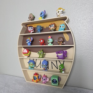 May include: A beige, honey pot-shaped shelf displaying a collection of colorful character figurines. The shelf has multiple levels and the word "HONEY" is written on the front. The figurines include characters from various animated movies and shows.