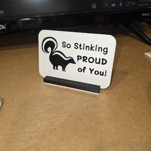 May include: A white rectangular sign with rounded corners, featuring a black skunk illustration and the text "So Stinking PROUD of You!". The sign is held by a black rectangular base, suitable for display on a desk or shelf.