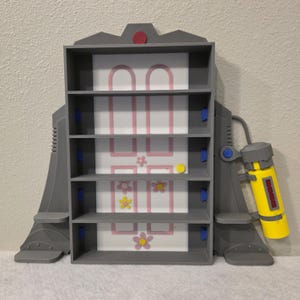 May include: A gray, multi-shelf storage unit with a white and pink door design, adorned with flower accents. A yellow and gray toy is attached to the side. The unit has four shelves and a red circle on top.