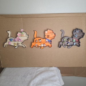 May include: Three wooden cat-shaped shelves, each with multiple levels, are filled with small character figurines. The shelves are painted in cream, orange, and gray, and are mounted on a cardboard background. The figurines are of various colors and designs.
