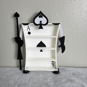May include: A white Ace of Spades-shaped shelf with three shelves. The shelf has black spade symbols and the letter "A" on it. A black staff and arm are attached to the shelf, creating a whimsical design.