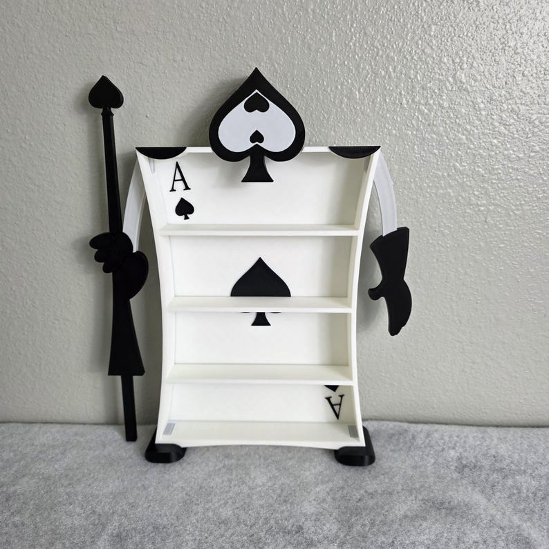 May include: A white Ace of Spades-shaped shelf with three shelves. The shelf has black spade symbols and the letter "A" on it. A black staff and arm are attached to the shelf, creating a whimsical design.