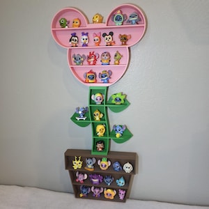 May include: A pink Mickey Mouse-shaped shelf with multiple levels, holding various character figurines. The shelf is mounted on a wall, with a green stem and brown pot-shaped base, also holding figurines. The figurines are of various colors and designs.
