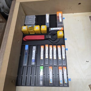 May include: A wooden drawer filled with various woodworking tools and accessories. The tools include a red adjustable wrench, yellow and clear plastic containers, and several drill bits in clear plastic cases. The tools are organized in a black tray.