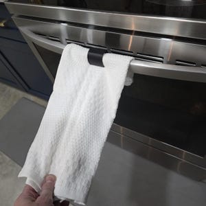 Könnte beinhalten: Ein weißes Küchentuch mit Waffelstruktur hängt an einem Edelstahl-Backofen. Das Handtuch wird von einem schwarzen Clip gehalten. Der Ofen hat eine schwarze Glastür und einen Edelstahlgriff.