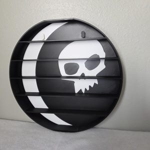 May include: A black and white circular display shelf featuring a skull design. The shelf has multiple horizontal slots for displaying items. The skull is white, and the background is black with a white stripe. The shelf is mounted on a white wall.