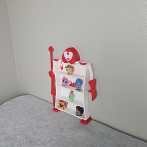 May include: A whimsical, red and white shelf shaped like the Ace of Hearts card. The shelf has multiple levels holding small character figurines. The top of the shelf features a heart design and the letter "A".