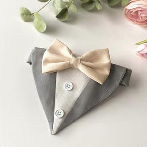 May include: A triangular pet bandana designed to resemble a tuxedo, featuring a grey fabric base with a central white panel, two white buttons, and a beige satin bow tie attached at the top. The accessory has a structured, formal appearance.