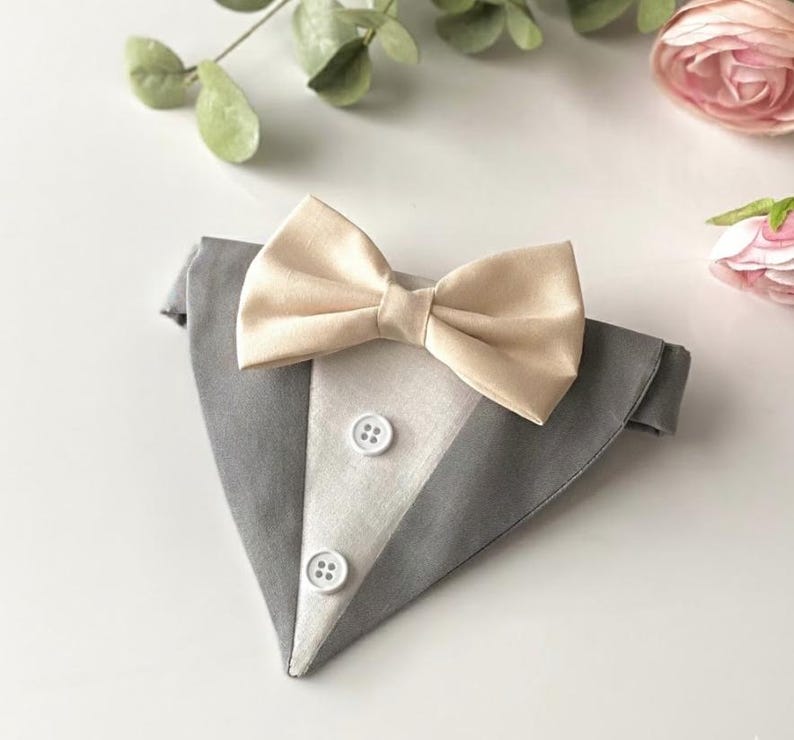 May include: A triangular pet bandana designed to resemble a tuxedo, featuring a grey fabric base with a central white panel, two white buttons, and a beige satin bow tie attached at the top. The accessory has a structured, formal appearance.