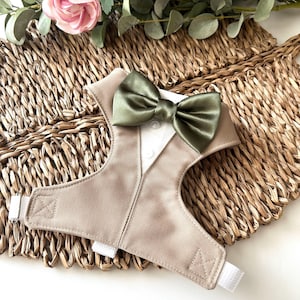 May include: A tan dog harness designed to resemble a tuxedo. It has a white shirt front, a khaki-coloured waistcoat, and an olive green bow tie. The harness has adjustable straps and a hook and loop fastening.