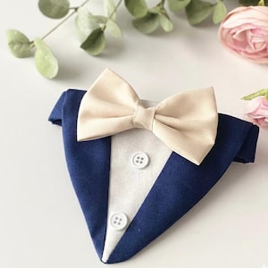 May include: A triangular pet bandana designed to resemble a tuxedo, featuring a dark navy blue fabric base with a white center panel, two white buttons, and a cream-colored fabric bow tie attached at the top center of the garment.