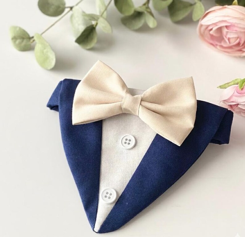 May include: A triangular pet bandana designed to resemble a tuxedo, featuring a dark navy blue fabric base with a white center panel, two white buttons, and a cream-colored fabric bow tie attached at the top center of the garment.