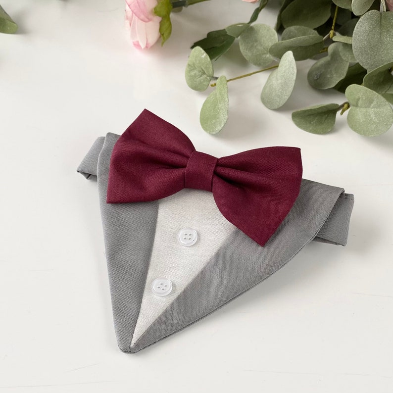 Puede incluir: Un pa&ntilde;uelo triangular para mascotas con dise&ntilde;o de esmoquin, que presenta una tela exterior gris, un panel central blanco con dos botones peque&ntilde;os blancos y una pajarita de color burdeos oscuro unida al borde superior.